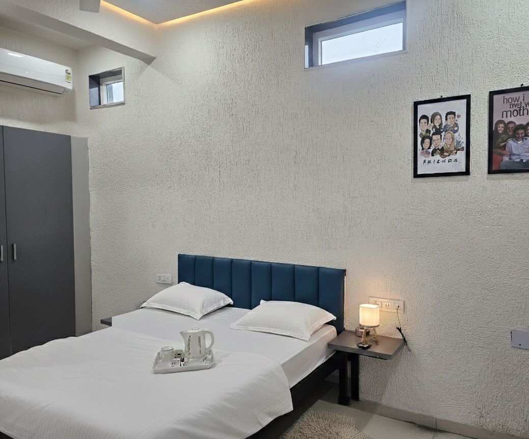 A bedroom with a bed, bedside table with a lamp, a small window, and a wall-mounted air conditioner.