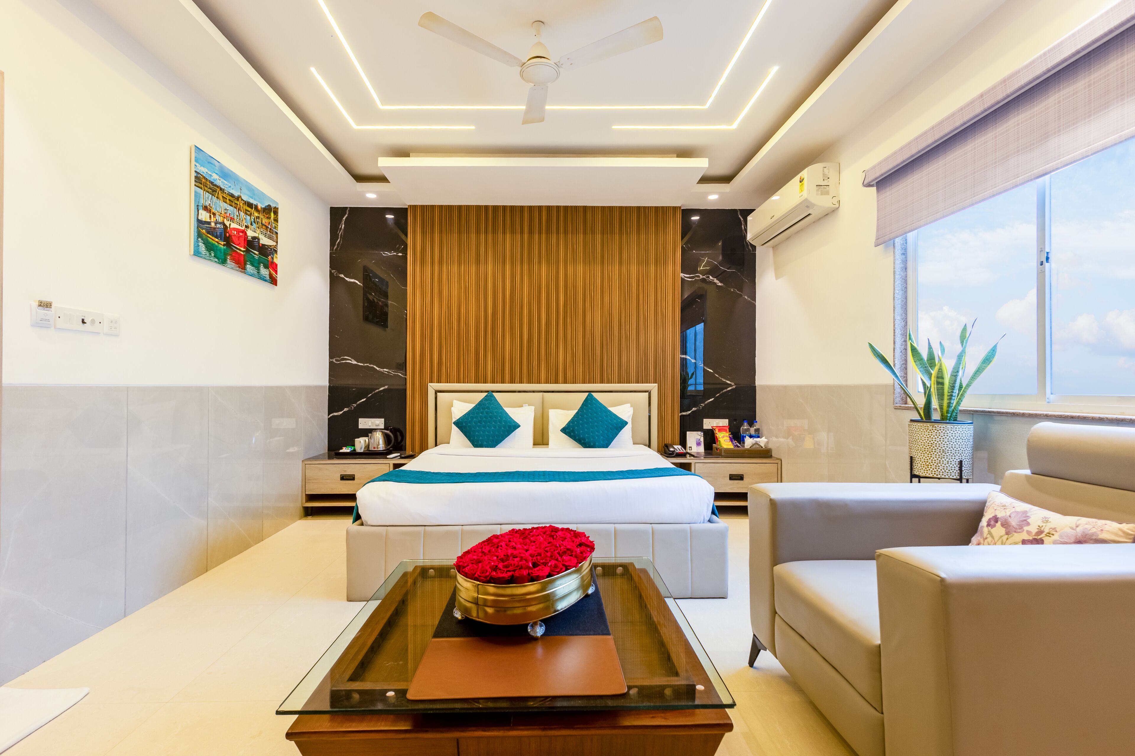 A modern hotel room with a large bed, a glass coffee table with a red flower centerpiece, a sofa, and a view of the sky through the window.