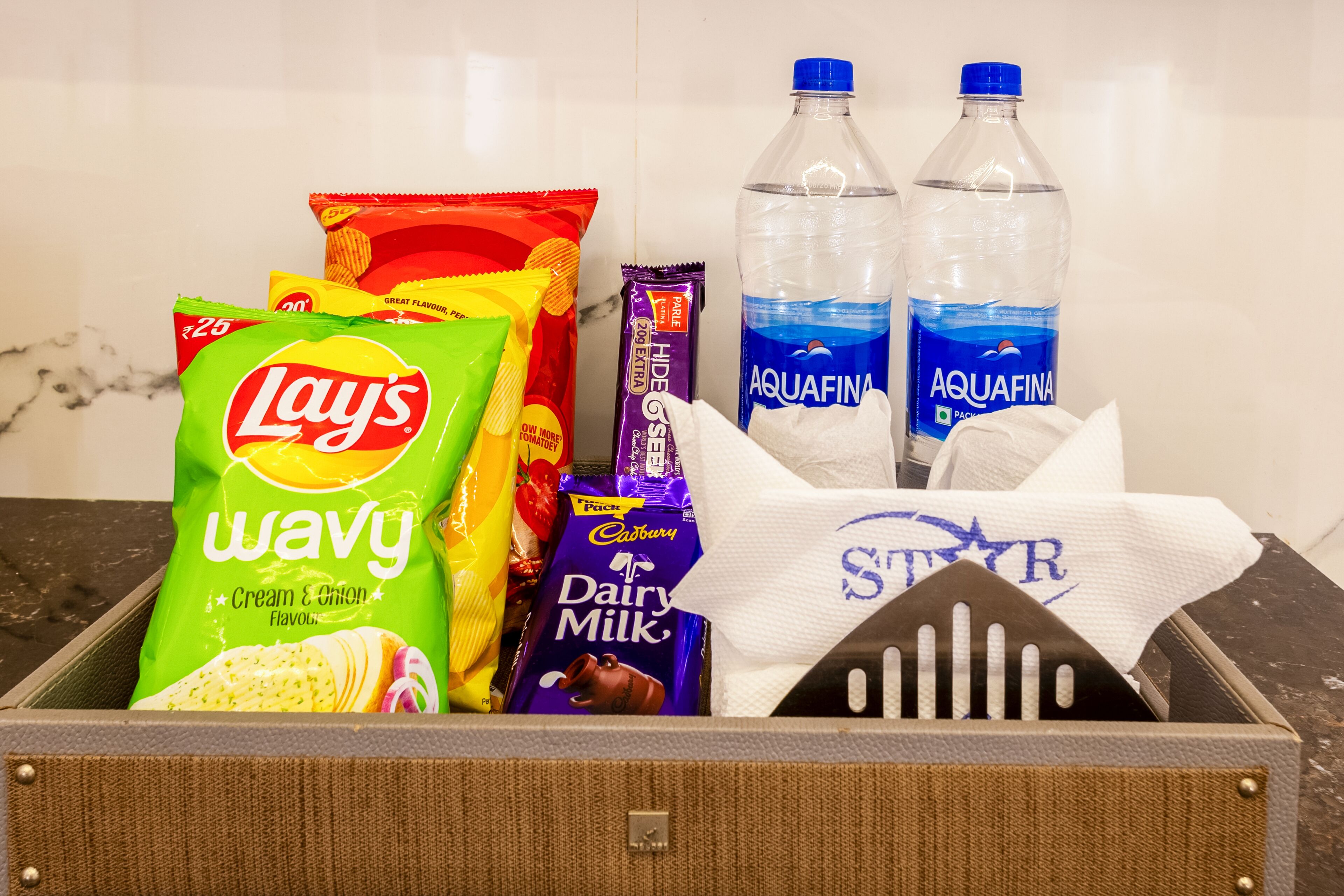Un cajón con aperitivos que contiene papas fritas Lay's Wavy, galletas saladas Face, chocolate Cadbury Dairy Milk y botellas de agua Aquafina.