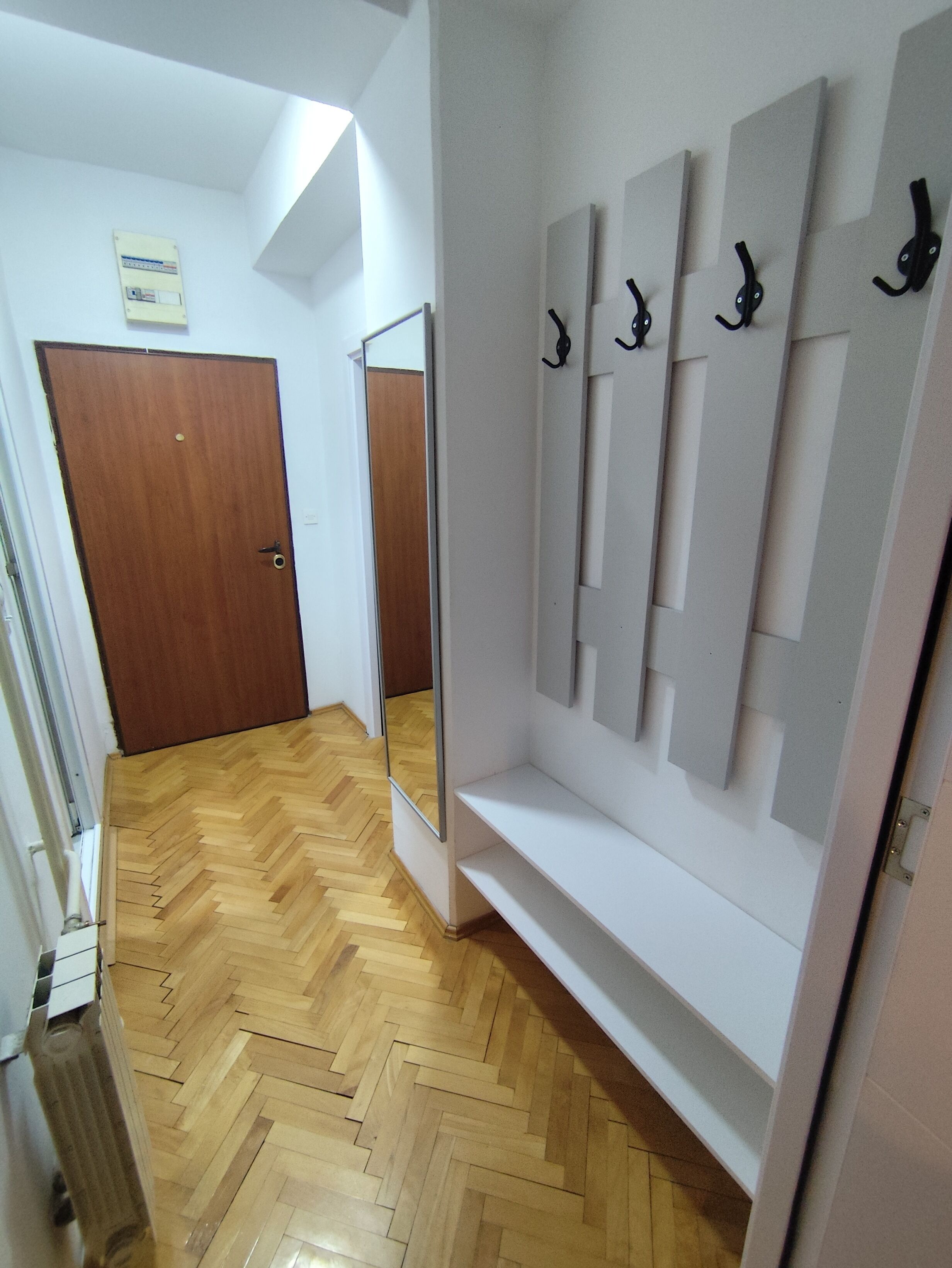 Um corredor com piso de madeira em padrão herringbone, uma porta de madeira e uma parede branca com espelho e ganchos.