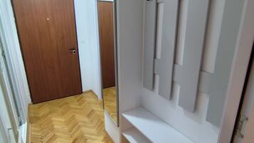 Um corredor com piso de madeira em padrão herringbone, uma porta de madeira e uma parede branca com espelho e ganchos.