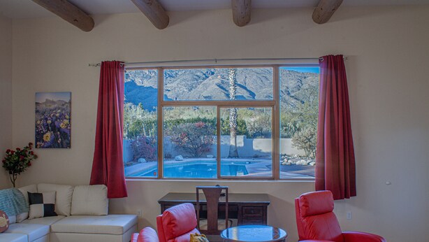 A living room with a large window, a sofa, two red chairs, a small table, and a painting on the wall. - 1 Acre Estate Home,Mtn. Views,Pool,Billiards,Private Desert:Saguaro Sanctuary@Catalina Foothills (Tucson)