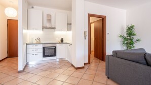 Een moderne keuken met witte kastjes, een inbouwoven en een aanrecht. In de woonkamer staat een grijze bank en een groene plant.