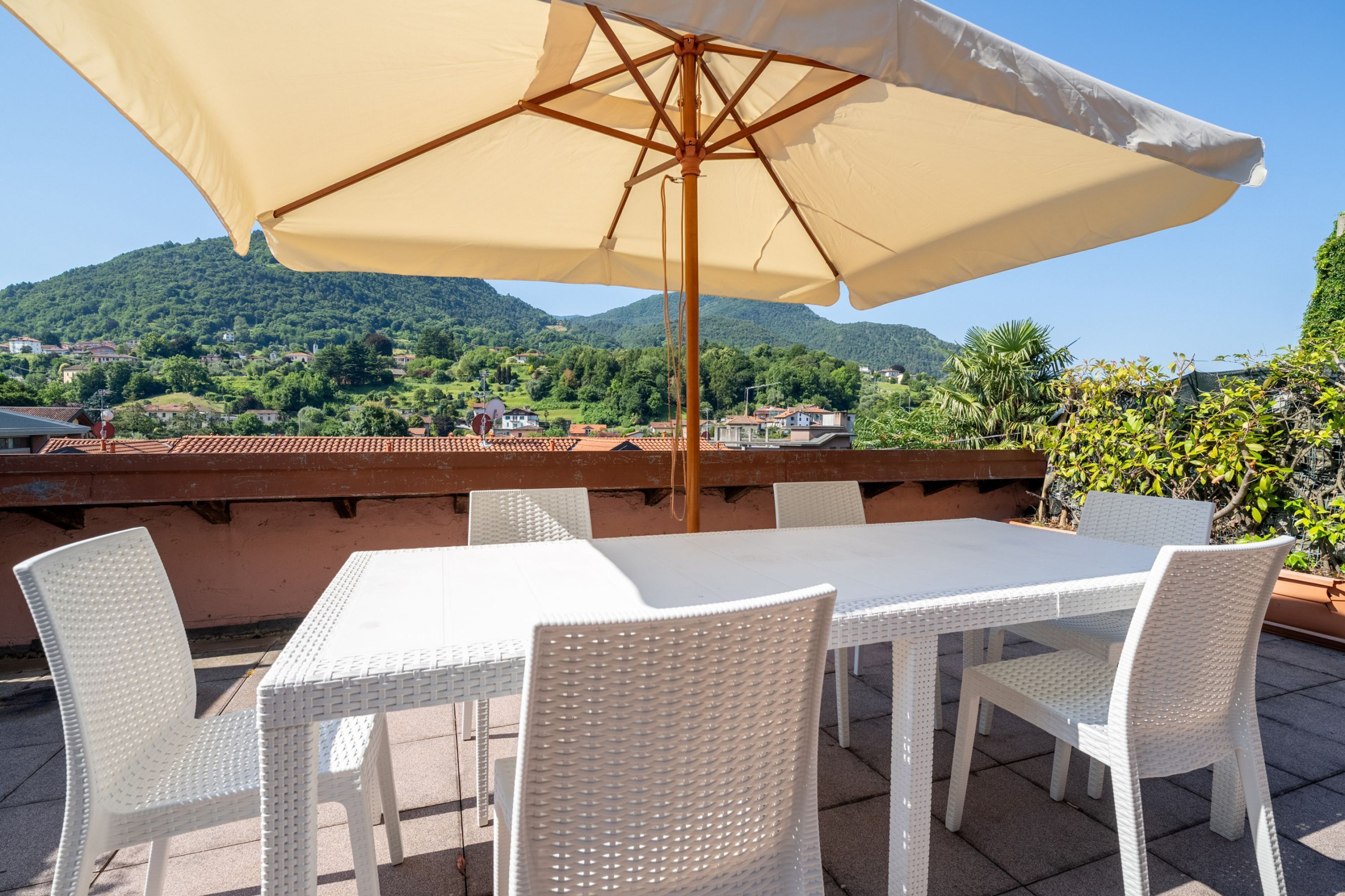 A terrace with a table and chairs, a large umbrella, and a scenic view of green hills and houses.