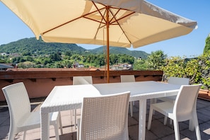 A terrace with a table and chairs, a large umbrella, and a scenic view of green hills and houses. - Cascine Trilocale con Terrazza by Wonderful Italy (Bellagio)