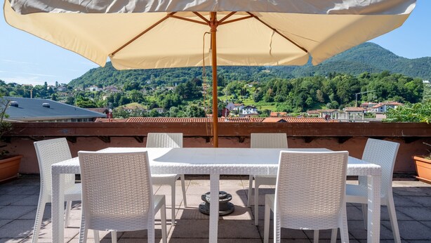 Een terras met een tafel en stoelen onder een grote parasol, met uitzicht op een bergachtig landschap.