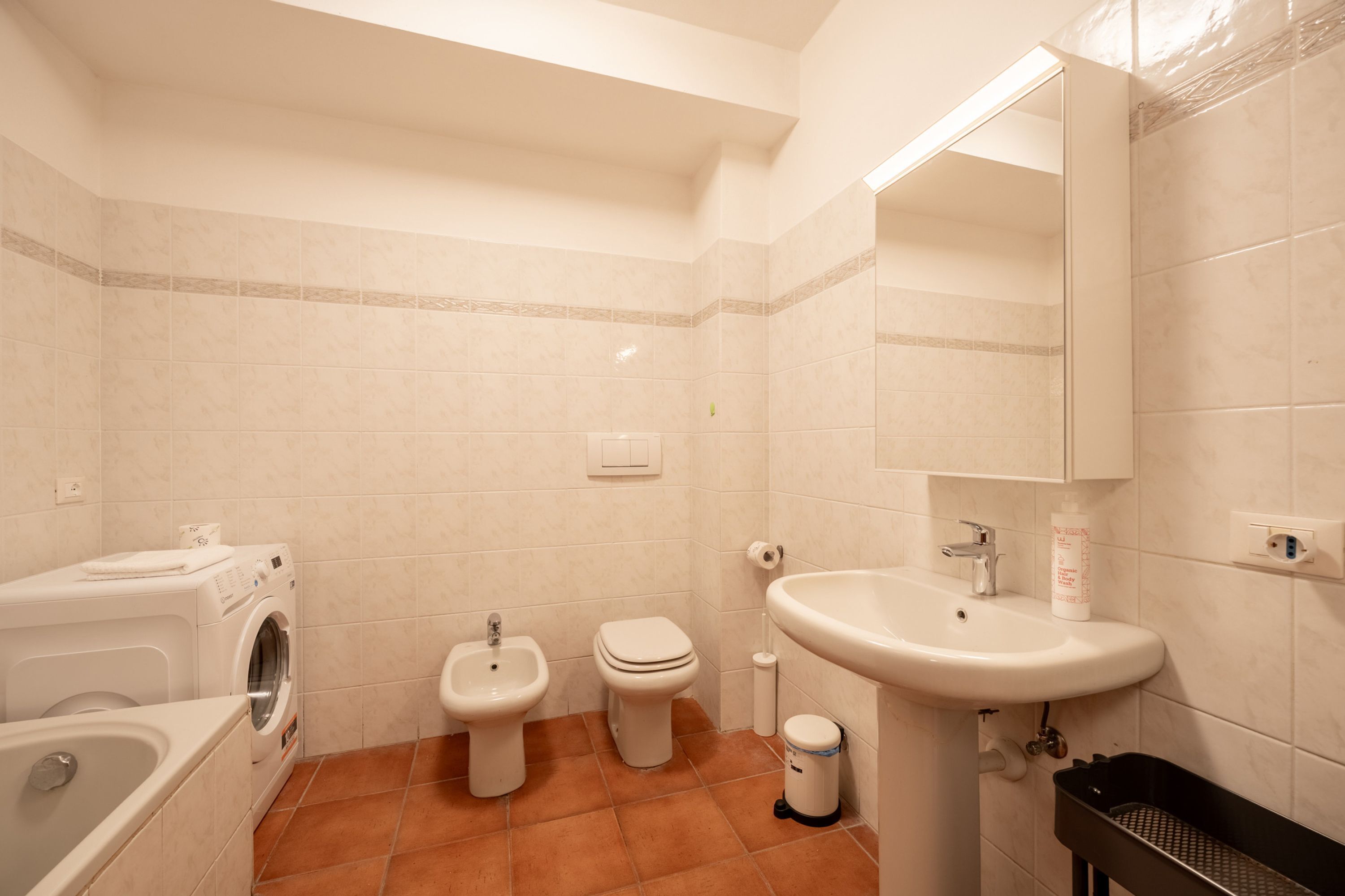 A bathroom with a washing machine, two toilets, a bidet, a sink, and a trash bin.