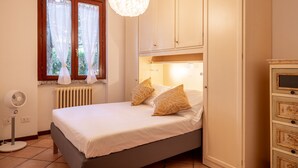 Een slaapkamer met een bed, een radiator, een raam met gordijnen, een ventilator en een kledingkast.