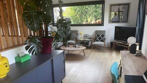 A modern living room with a wooden floor, a large potted plant, a sofa, a coffee table, a TV, and a laptop on a desk. - 150 m² wooden family house with 3 terraces, close to the sea and surf spots! (Landunvez)