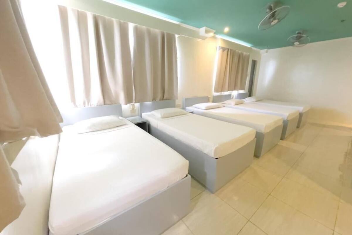 A hotel room with three single beds, a large window with curtains, and a teal-colored ceiling.