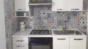 A compact kitchen with white cabinets, a gas stove, a built-in oven, and a sink. Decorative tiles adorn the backsplash, and there are two small figurines on the upper cabinets. - The Carusians rent short (Marsala)
