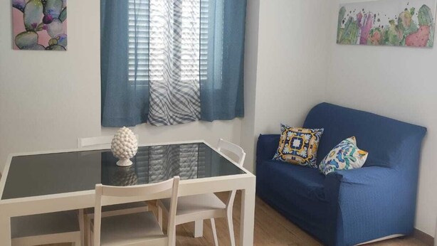 A room with a dining table, chairs, a blue sofa with patterned cushions, a window with blue curtains, and two wall art pieces. - The Carusians rent short (Marsala)