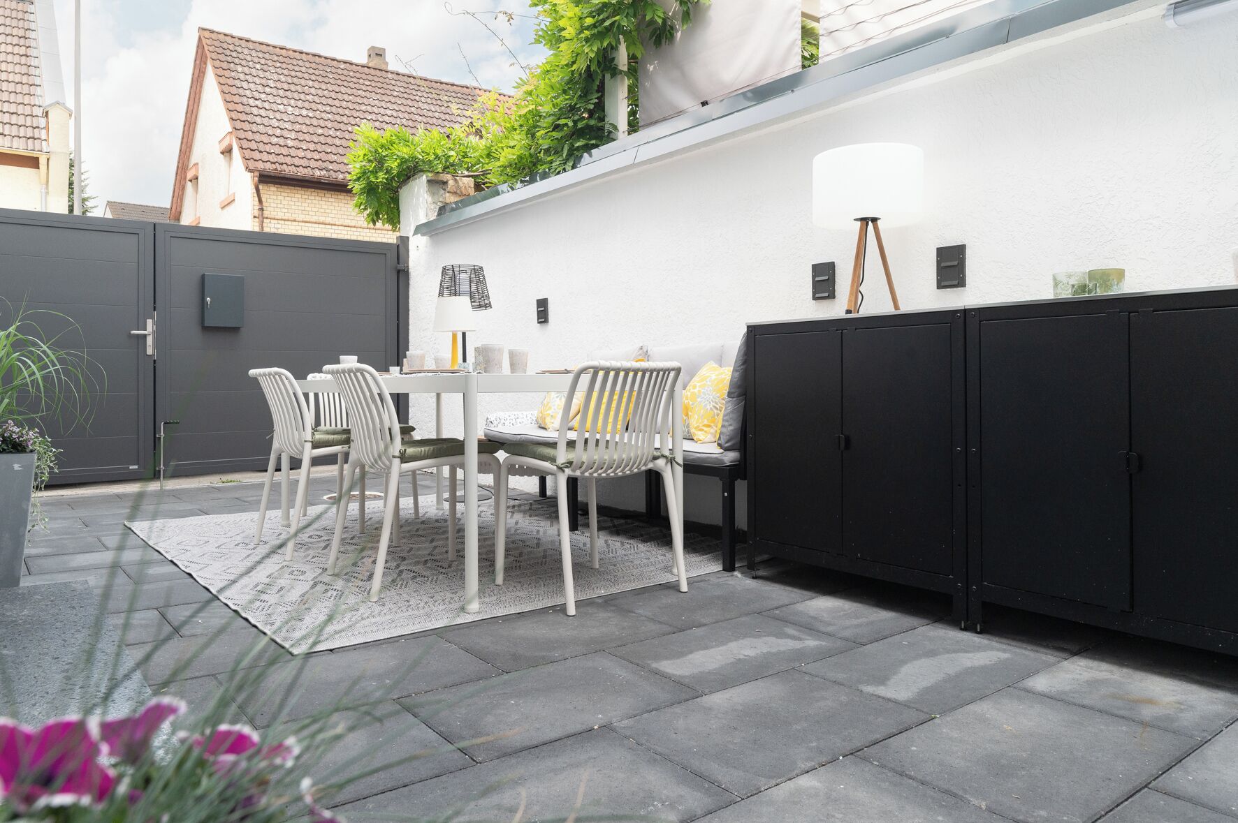 Eine Terrasse mit Tisch und Stühlen, einem schwarzen Stauraum und einer weißen Wand mit einem Fenster.
