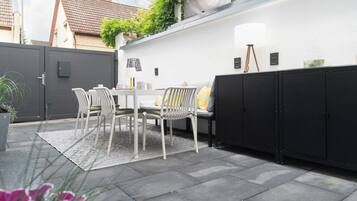 Eine Terrasse mit Tisch und Stühlen, einem schwarzen Stauraum und einer weißen Wand mit einem Fenster.