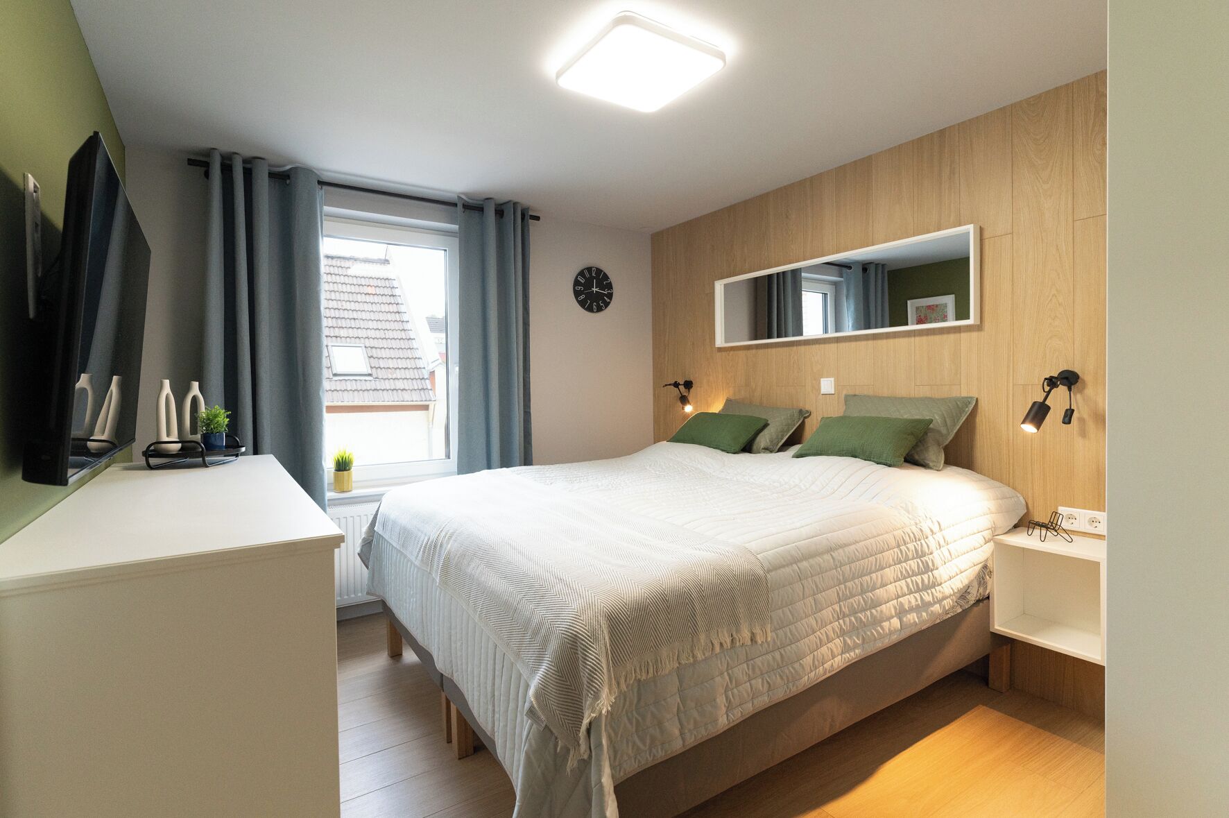 Een slaapkamer met een groot bed, nachtkastjes, een spiegel en een raam met gordijnen.
