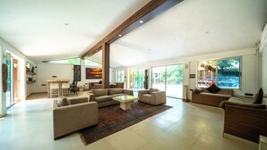 A spacious living room with a large sofa set, a coffee table, and a rug. There is a dining area with a table and chairs, and a kitchen in the background. - The Amore Collective (Bengaluru)