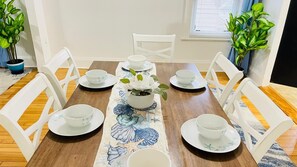 Une table à manger dressée avec de l’assiette et des bols blancs, un chemin de table à motifs floraux et des plantes en pot de chaque côté.