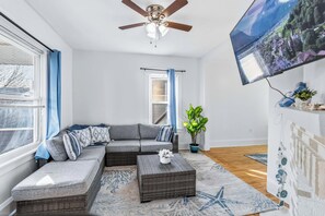 Un salon moderne avec un canapé d’angle, un téléviseur à écran plat, un ventilateur de plafond et une grande fenêtre équipée de stores.