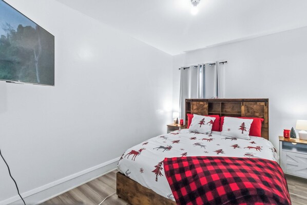 Une chambre à coucher comprenant un lit, une tête de lit en bois, une table de chevet, un téléviseur fixé au mur et une fenêtre avec des rideaux.