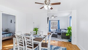 Une pièce moderne et bien éclairée, avec une table à manger dressée pour quatre personnes, un ventilateur de plafond et une vue sur un espace de vie comprenant un canapé et une grande fenêtre.