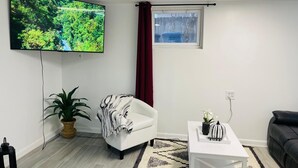 Ein modernes Wohnzimmer mit einem Flachbildfernseher, einem weißen Sessel, einem kleinen Tisch mit Dekoration, einer Topfpflanze und einem grauen Sofa.