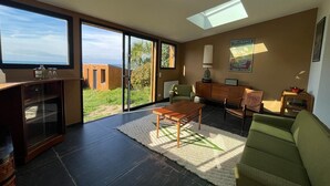 A modern living room with a large glass door leading to a balcony, a wooden coffee table, a green sofa, and a painting on the wall. - Seaside Chalet with Direct Water Access (Fermanville)