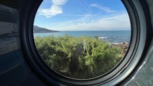 A round window with a view of a coastal landscape, including trees and the sea. - Seaside Chalet with Direct Water Access (Fermanville)