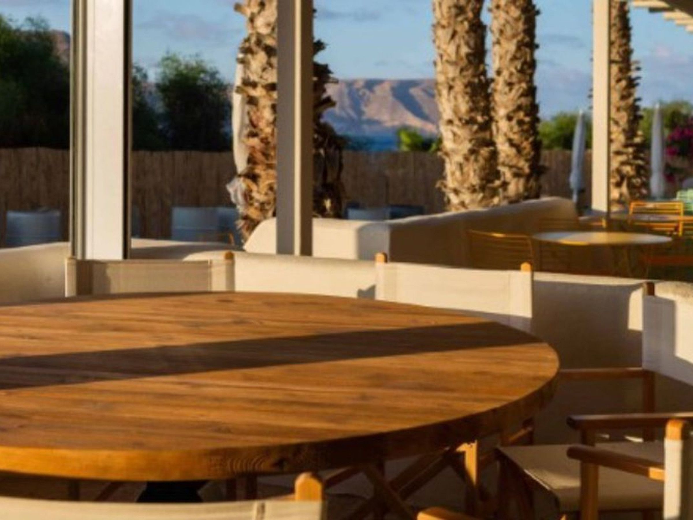 A round wooden table with white cushioned chairs in a room with large windows and a view of palm trees and mountains.