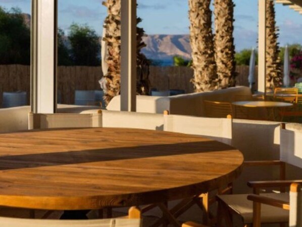 A round wooden table with white cushioned chairs in a room with large windows and a view of palm trees and mountains.