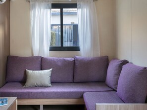 A purple sofa with a cushion, a window with white curtains, and a view of the outside.