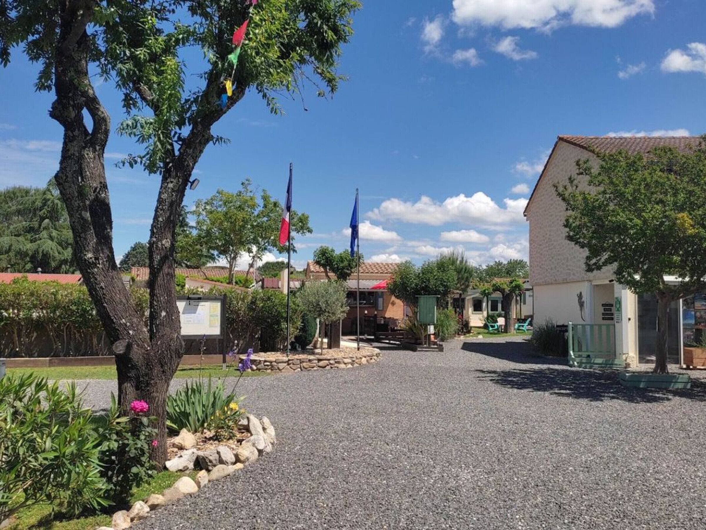 A gravel parking area with a building on the right and a tree with colorful flags on the left.