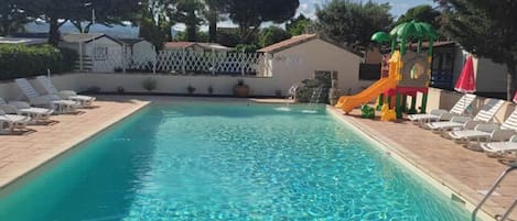 A rectangular swimming pool with clear blue water, surrounded by white lounge chairs and a children's play area with a slide.