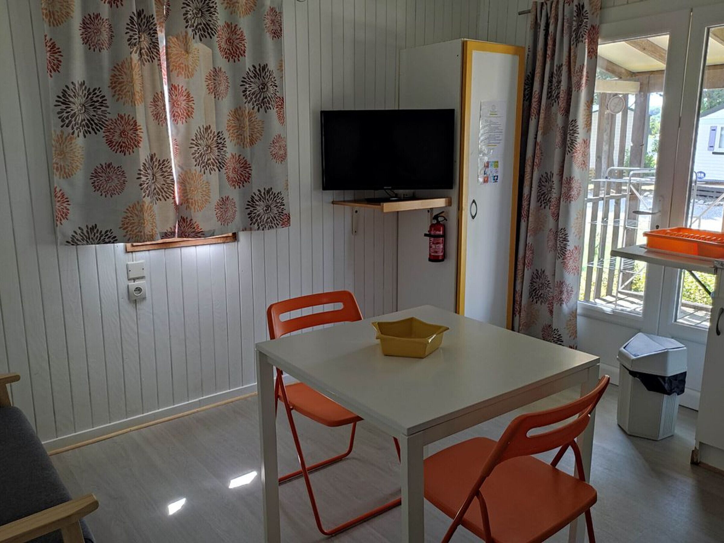 Una habitació amb taula blanca, cadires taronges, televisió, extintor i finestra amb cortines.