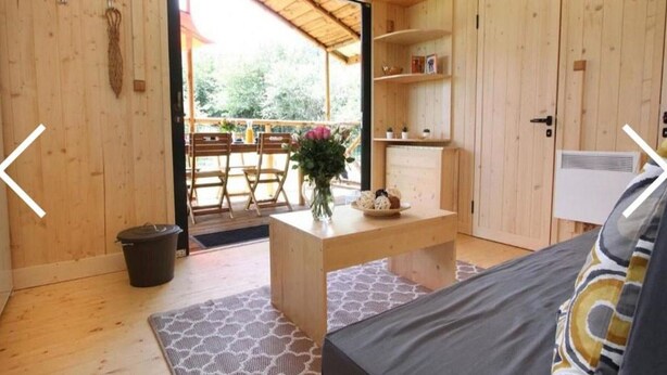 Une pièce en bois comprenant un lit, une petite table avec un vase, une poubelle et une porte donnant sur un espace extérieur avec une table et des chaises.