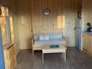 Quarto compacto com piso de madeira, um pequeno banco de madeira, um sofá branco, um relógio na parede e uma área de cozinha com geladeira e micro-ondas.