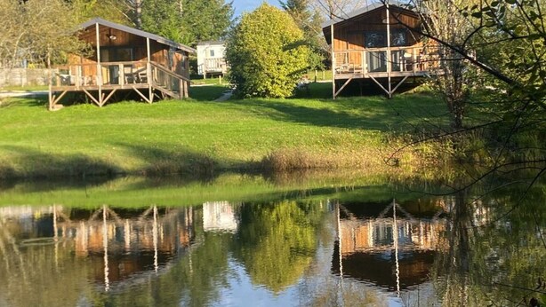 Deux cabanes en bois avec accès à une terrasse, entourées d’un étang et d’arbres.