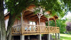 A wooden cabin with a covered porch, supported by concrete pillars, surrounded by trees and grass. - Cozy chalet with terrace in Naucelle - Idyllic location (Naucelle)