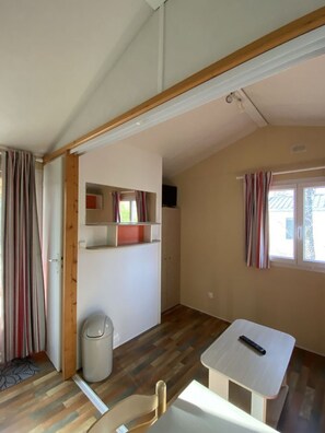 Una habitación con techo de madera, una mesa blanca con un control remoto, un cesto de basura y una ventana con cortinas.