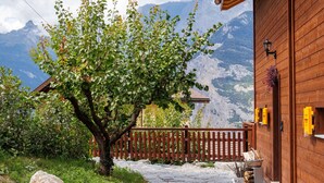 Ein Holzhaus mit einem Steinweg, der zu einer Holzdiele führt, umgeben von Grünpflanzen und Bergen im Hintergrund.