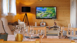 Ein gemütliches Holzhaus-Interieur mit einem Esstisch, der für sechs Personen gedeckt ist, einem an der Wand befestigten Fernseher und einer Stehlampe.