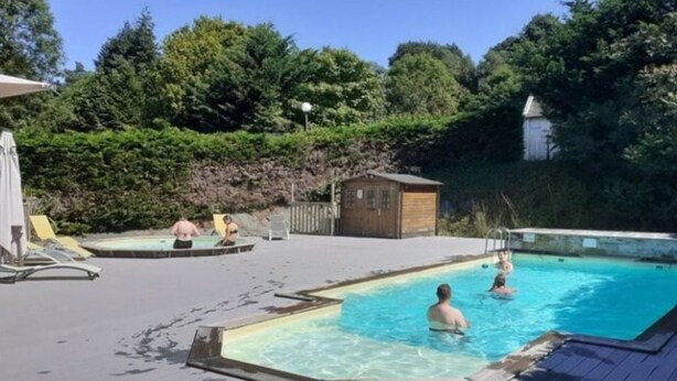 A swimming pool area with a wooden deck, a small wooden building, and lush greenery in the background. - Mobile Home with Terrace in Guégon (Guégon)