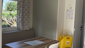 A compact room with a wooden table, two white chairs, a bench, a window with patterned curtains, a door with a notice, and a trash bin. - Mobile Home with Terrace in Guégon (Guégon)