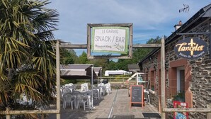 An outdoor seating area with tables and chairs, a sign for 'LEGARVIG SNACK / BAR', and a building with a sign 'Tancelot Bières de Bretagne'. - Mobile Home with Terrace in Guégon (Guégon)