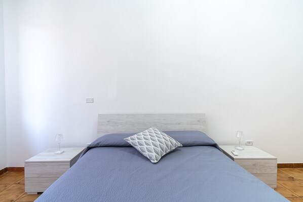 A bed with a grey bedspread and a patterned pillow, two bedside tables with lamps, and a plain white wall.