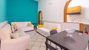 A living room with two white sofas, a dining table with a clear glass bowl, and a kitchen area with wooden cabinets and a tiled backsplash. - Apartment 'Ciuri' with Wi-Fi and Air Conditioning (Avola)