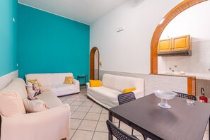 A living room with two white sofas, a dining table with a clear glass bowl, and a kitchen area with wooden cabinets and a tiled backsplash.