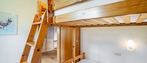 Una cama litera con escalera de madera, pared blanca y una lámpara.