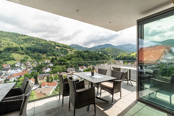 A balcony with wicker furniture, a table, and a glass door overlooking a town and mountains.