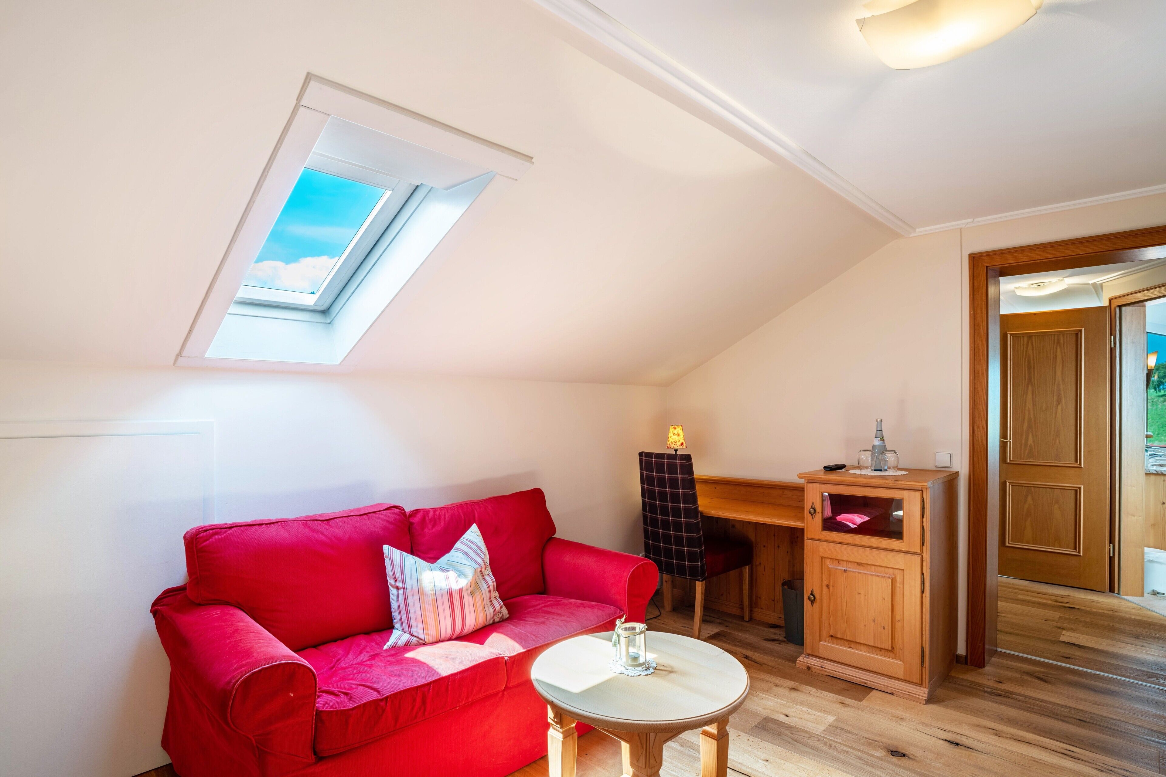 A red sofa, a small round table, a wooden cabinet, a desk, and a skylight in a room with wooden flooring.