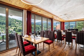 A restaurant with a large table and chairs, large windows with a view of a forested area, and a wooden ceiling.